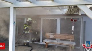 Zwei Feuerwehrleute sind draußen und spritzen Wasser. Ein Holzbank und eine Glastür sind in der Nähe.