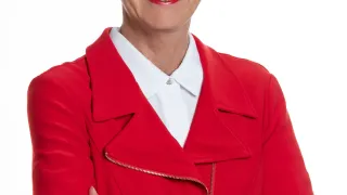 A woman with short blonde hair is wearing a red blazer and a white shirt. She is smiling and posing for a picture with her hands crossed.