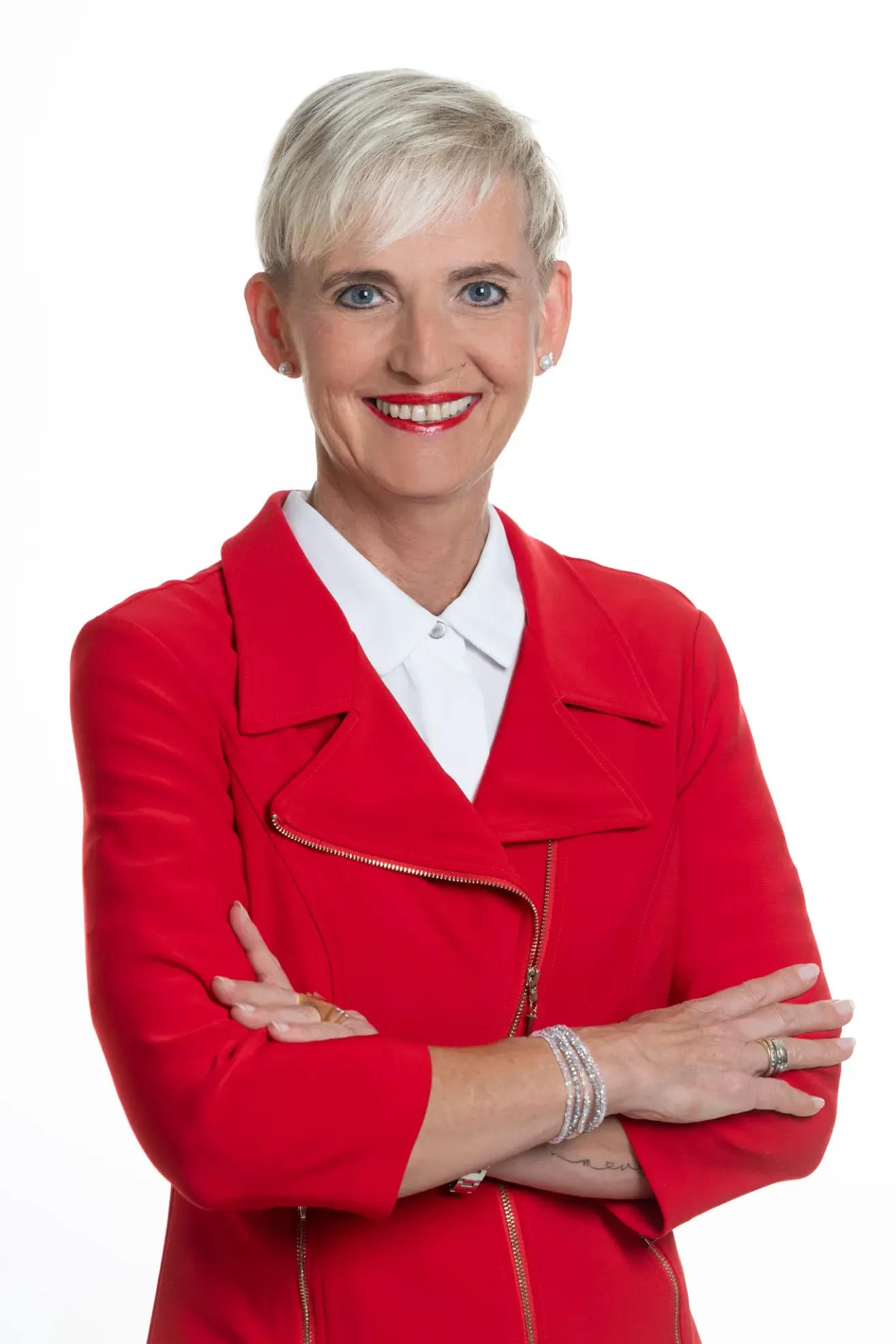 A woman with short blonde hair is wearing a red blazer and a white shirt. She is smiling and posing for a picture with her hands crossed.