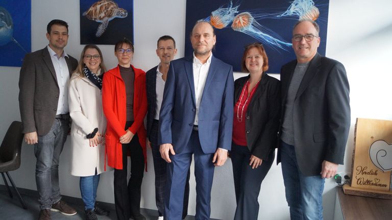 A group of people, five women and two men, stand in front of a wall with two large paintings. They are dressed in winter clothes and appear to be posing for a photograph.