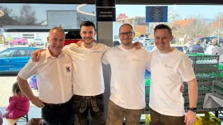 Four men in white shirts and brown shorts stand in a store, smiling for a photo with a glass window in the background.