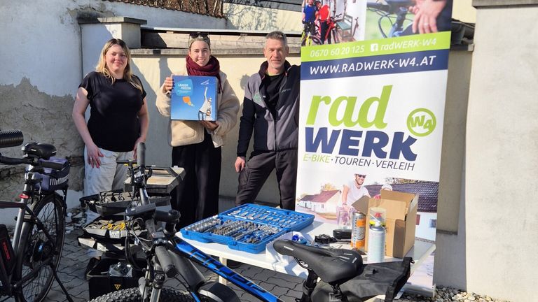 Drei Personen stehen neben einem Fahrrad mit einer Kiste und einem Werkzeugsatz auf einem Tisch. Eine hält eine Kiste mit der Aufschrift Leader Region. Ein Schild dahinter steht 'rad WER K'.