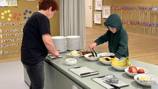 A woman and a child prepare food at a buffet. Various fruits and vegetables are on the table, with plates and utensils nearby. Behind them, a wall is decorated with colorful shapes and frames.