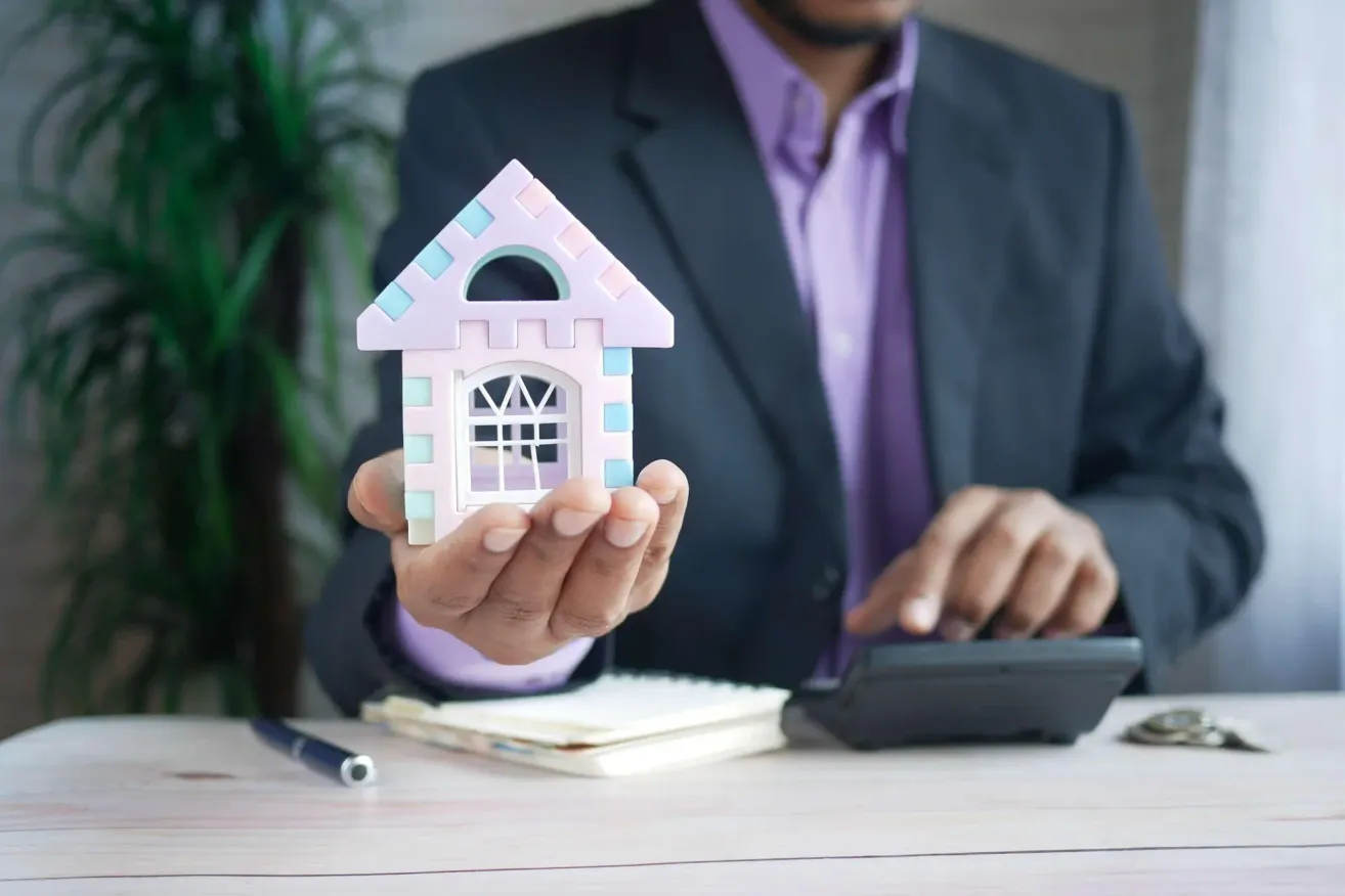 A man in a suit holds a small house model, possibly discussing real estate. A calculator and pen are on the desk.