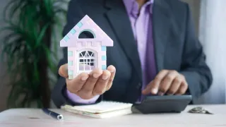 A man in a suit holds a small house model, possibly discussing real estate. A calculator and pen are on the desk.
