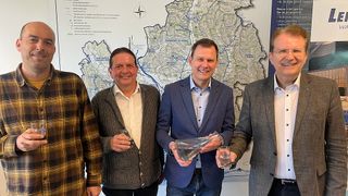 Four men stand in front of a map, smiling and holding glasses of water. The man on the left wears a plaid shirt. The middle man wears a gray sweater, and the right man wears a suit.