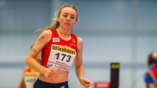 A female athlete with the number 173 on her jersey is running on a track, possibly in a competition. She has tattoos on her arms and is wearing a necklace and bracelets.