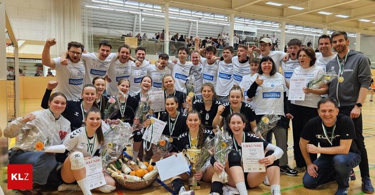 A team of athletes celebrates a victory with trophies, medals, and bouquets of flowers in a sports hall. Some are seated, and others are standing. They all wear sports uniforms.