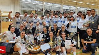 A team of athletes celebrates a victory with trophies, medals, and bouquets of flowers in a sports hall. Some are seated, and others are standing. They all wear sports uniforms.