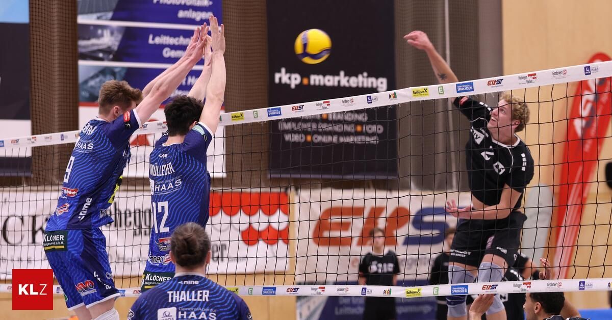 Volleyball players compete indoors. Two players block, while another spikes the ball. The net is marked with sponsors. Audience and advertisement banners are visible.