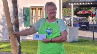 An older woman in a green apron stands next to a tree, holding a paper with a logo. Behind her is a building with green shutters and a car parked nearby.
