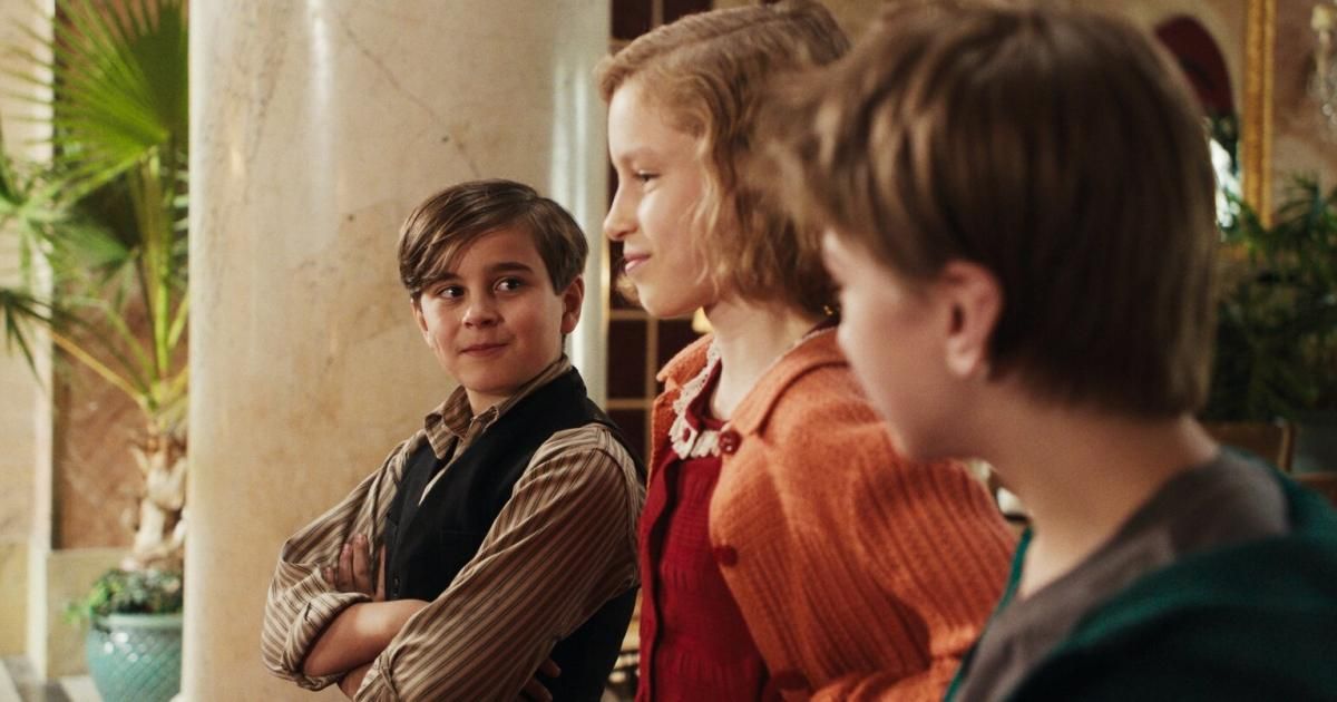 Three children are standing in a row. A boy on the left folds his arms, while a girl in the middle looks at him. Another child on the right stares ahead. Behind them, a marble wall and a wooden door are visible.