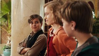 Three children are standing in a row. A boy on the left folds his arms, while a girl in the middle looks at him. Another child on the right stares ahead. Behind them, a marble wall and a wooden door are visible.