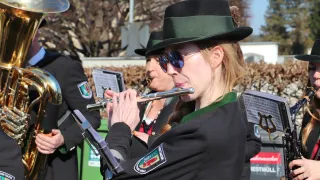 A woman in a black uniform plays a flute while another person holds a clarinet. They are standing outdoors, possibly in a park.