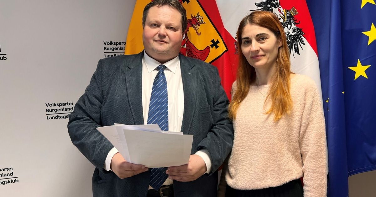 A man and woman stand next to each other holding papers, smiling in front of a flag with a dragon emblem.