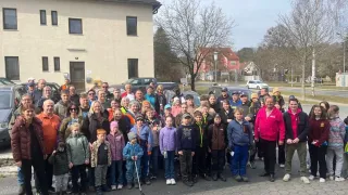A group of people, including adults and children, are posing for a picture in front of a building, with cars parked nearby and a streetlight visible in the background.