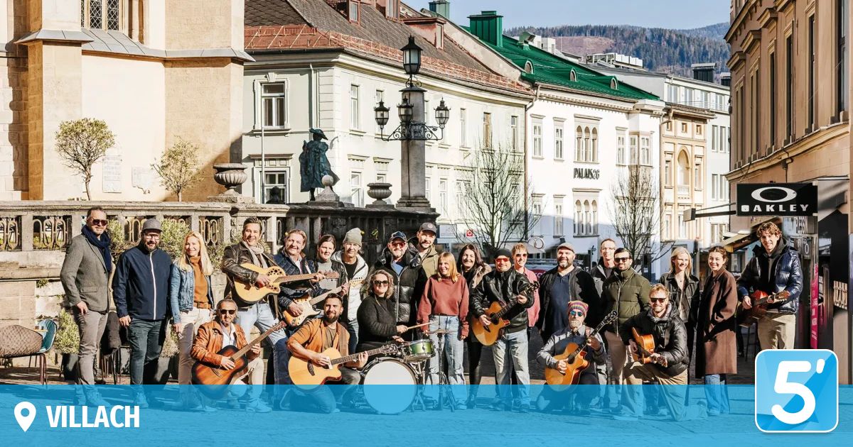 A group of musicians with guitars and a drum pose in front of an old building. Buildings with green roofs and a statue are in the background.