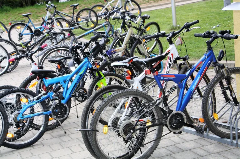 Viele Fahrräder sind auf einer gepflasterten Oberfläche mit Gras und einem Pfosten im Hintergrund geparkt. Die Fahrräder variieren in Farbe, Größe und Stil, und einige haben Wasserflaschen befestigt.