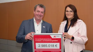 A man and woman hold a framed certificate for 2,985.018 euros, standing against a brown wall. Both are smiling and dressed formally. The man wears a blue suit, and the woman wears a pink blazer.