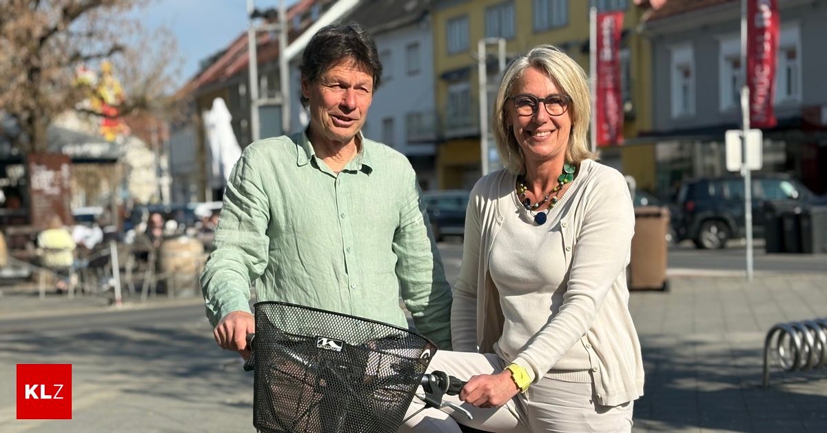 A man and a woman are smiling while standing next to a bicycle with a basket on a street. They are both dressed casually. The background shows buildings, parked cars, and street lights.