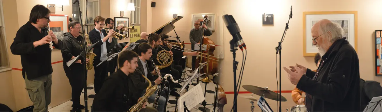 A group of musicians in a room, playing instruments like saxophones, trumpets, and trombones, with a pianist and a bass player. They are performing in a music studio with a piano, microphones, and a speaker.