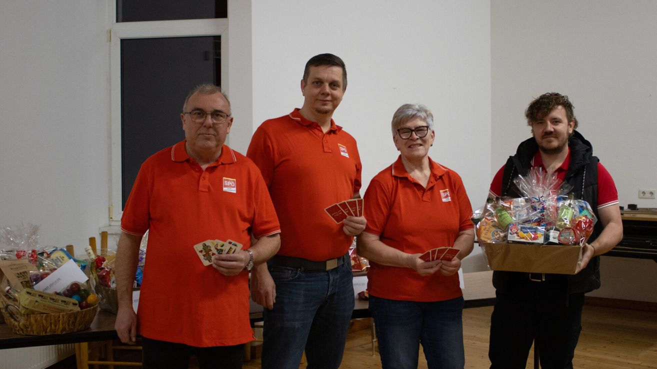 Vier Personen in orangefarbenen Hemden stehen in einem Raum zusammen. Die beiden Männer links und rechts halten jeweils ein Kartenset. Die beiden Frauen in der Mitte lächeln. Hinter ihnen steht ein Tisch.