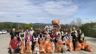 A group of people, including adults and children, poses for a photo in a parking lot, holding bags of trash. The background features a cloudy sky, trees, and mountains.