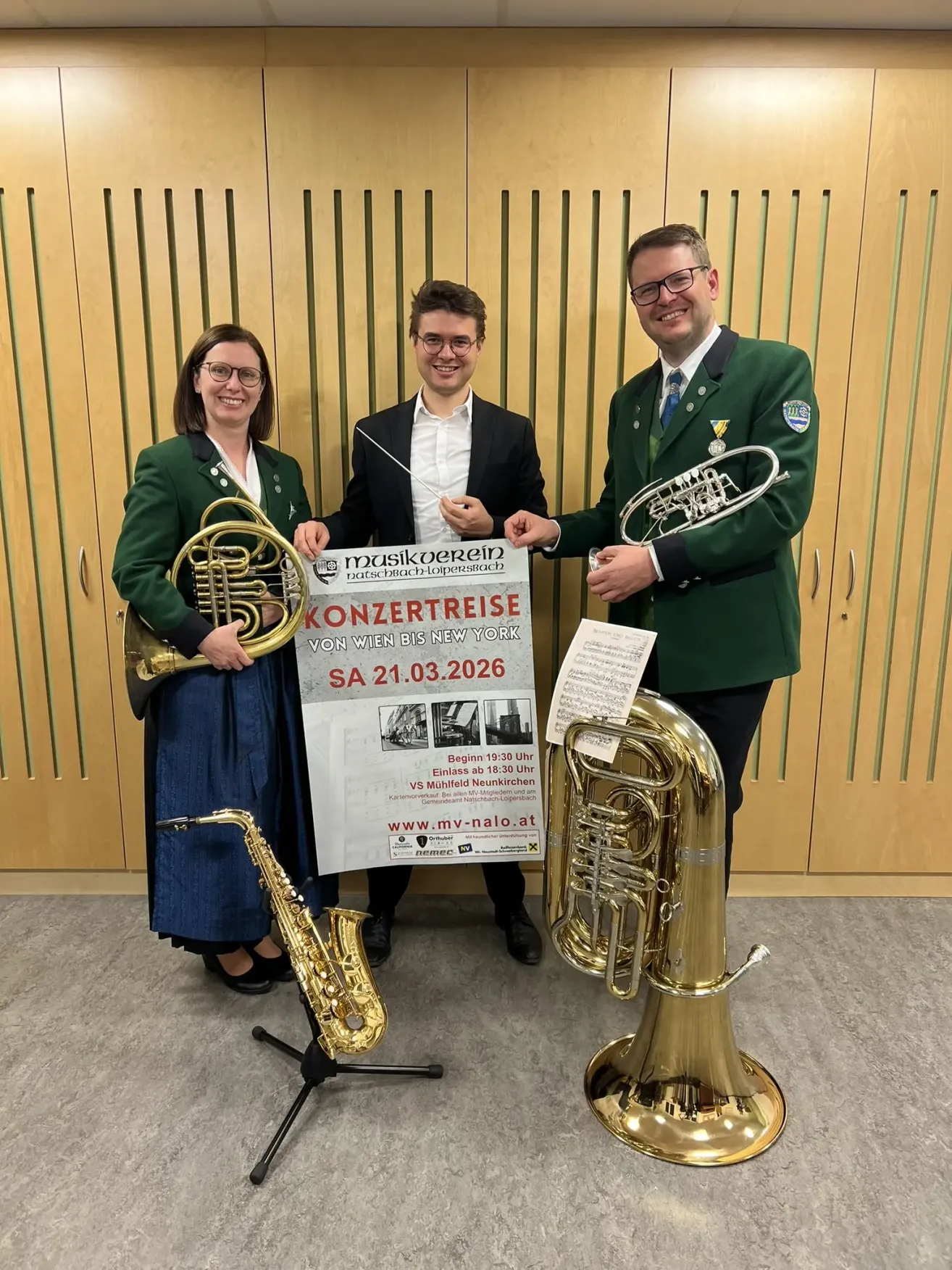 Three musicians pose with their instruments, smiling for a photo. They stand behind a banner promoting a concert tour. The man on the right holds a trumpet and a music sheet.