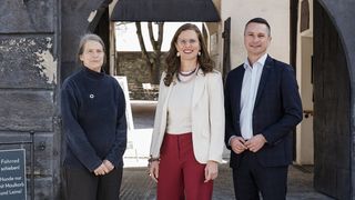 Drei Personen stehen vor einem Torbogen, eine Frau mit Brille und einer Halskette in der Mitte. Sie scheinen für ein Foto zu posieren.