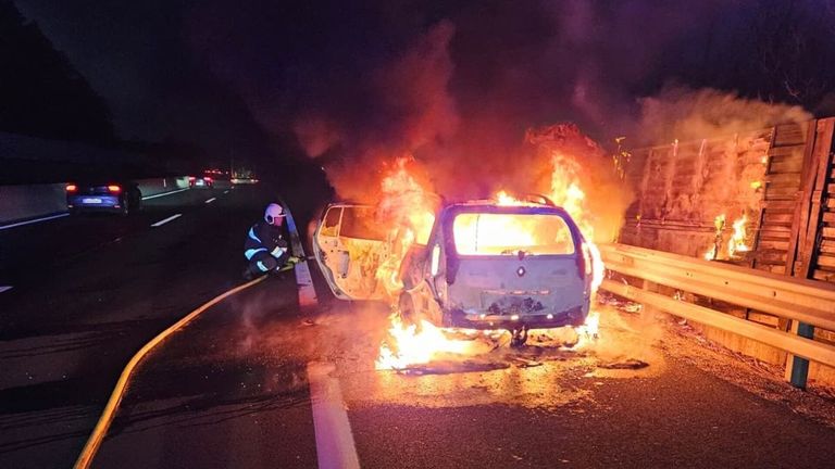 Ein Auto brennt nachts am Straßenrand. Ein Feuerwehrmann benutzt einen Schlauch, um die Flammen zu löschen.
