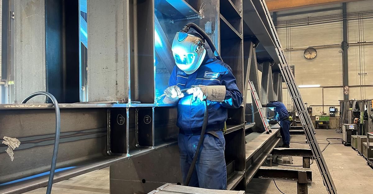 A man wearing a welding mask and a blue jumpsuit is welding metal in a factory. Another man is working on a metal structure in the background. The structure has labeled sections.