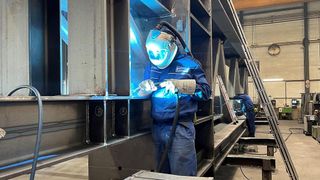 Ein Mann in einem Schweißerhelm und einer blauen Overall schweißt Metall in einer Fabrik. Ein weiterer Mann arbeitet im Hintergrund an einer Metallstruktur. Die Struktur hat beschriftete Abschnitte.