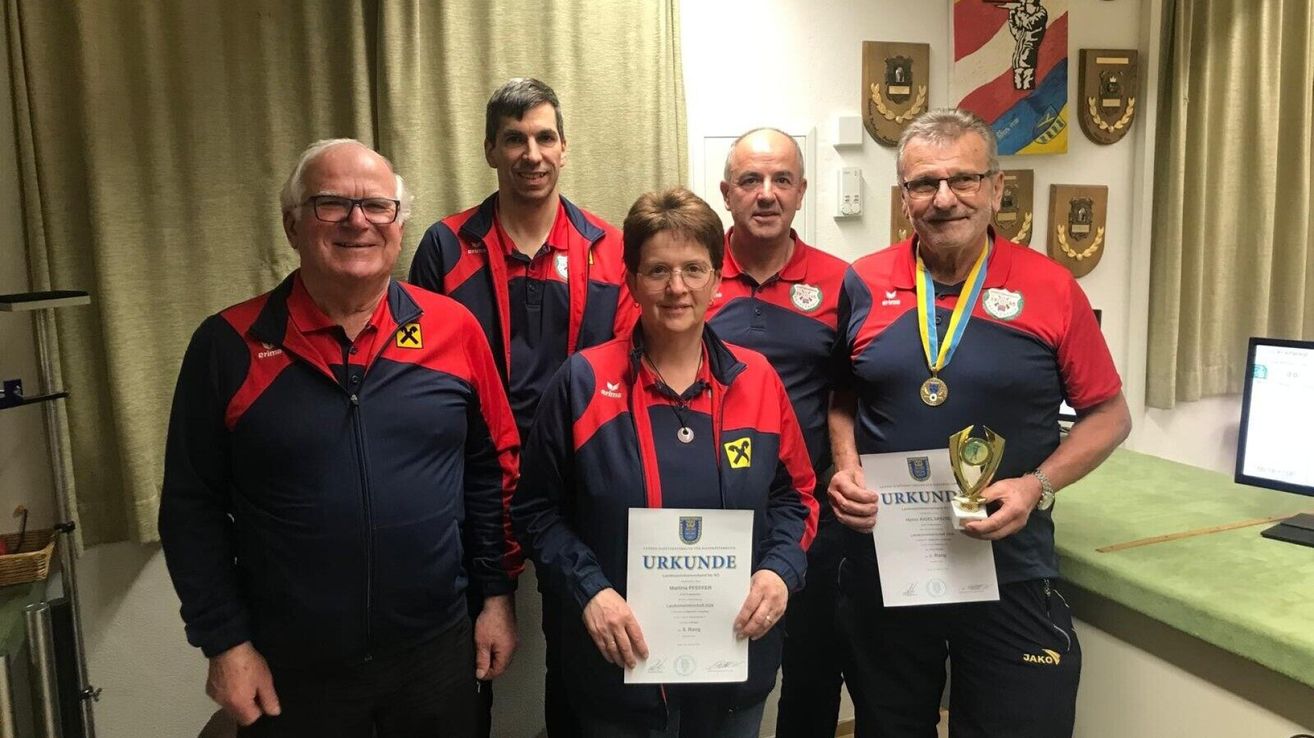 Fünf Menschen lächeln für ein Foto, halten Urkunden und Medaillen. Die Frau in der Mitte trägt eine Brille und eine Halskette. Die Männer tragen rote und blaue Jacken mit Logos und Medaillen. Ein Vorhang und Wanddekorationen sind im Hintergrund.