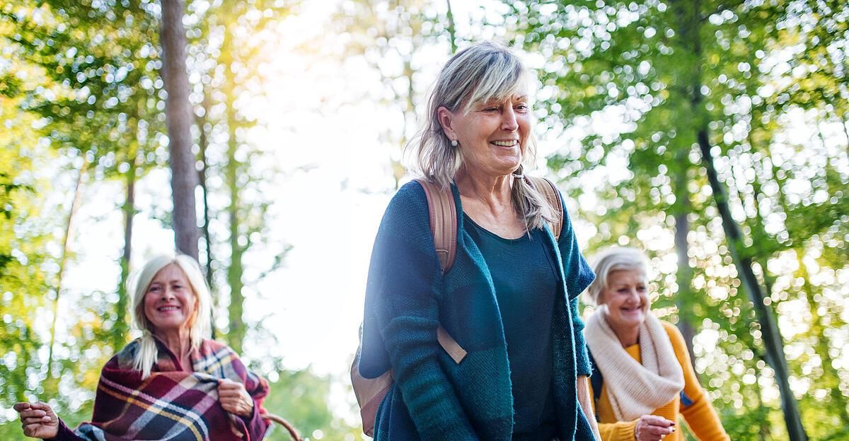 Drei ältere Frauen lächeln und gehen gemeinsam in einem sonnigen Wald, mit Bäumen im Hintergrund.