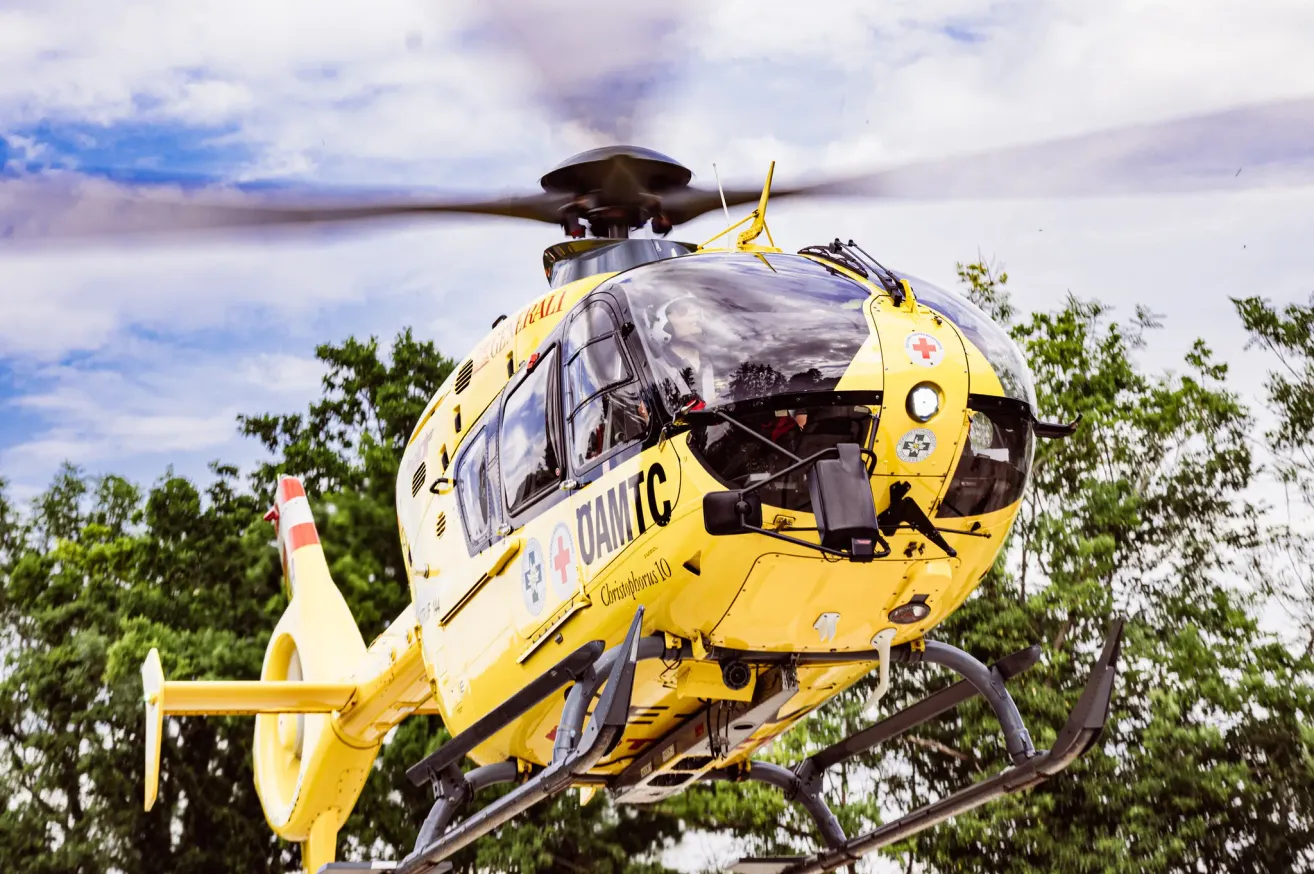 Ein gelber Hubschrauber mit 'OAMTC' und rotem Kreuz-Logo fliegt über Bäumen. Er hat eine Glaswindschutzscheibe und einen großen Propeller.