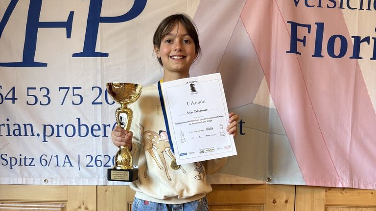 Ein junges Mädchen hält eine Trophäe und ein Zertifikat in den Händen, lächelt und posiert für ein Foto vor einem Banner.