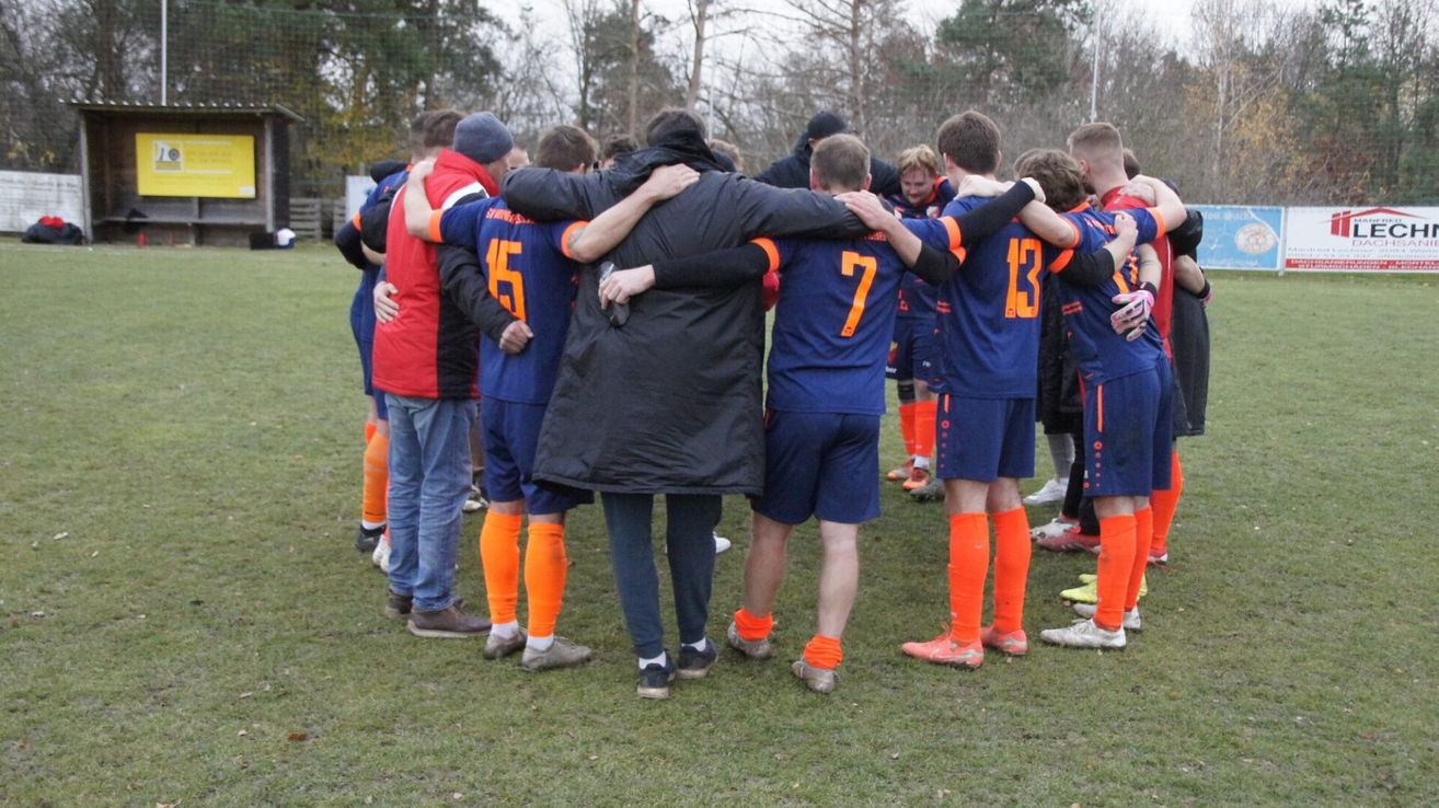 Eine Fußballmannschaft in blauen Trikots und orangefarbenen Socken versammelt sich auf einem Rasenfeld, mit Bäumen im Hintergrund.