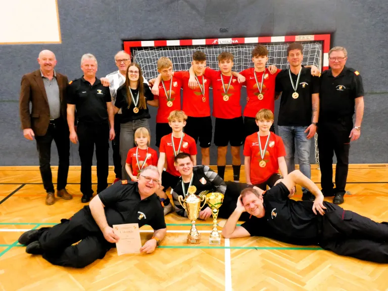 Eine Sportmannschaft mit roten Trikots und Medaillen posiert mit Trainern in einer Turnhalle. Ein Mann hält ein Zertifikat, während er auf dem Boden liegt. Zwei Pokale stehen zwischen zwei Männern. Ein Tor steht hinter ihnen.
