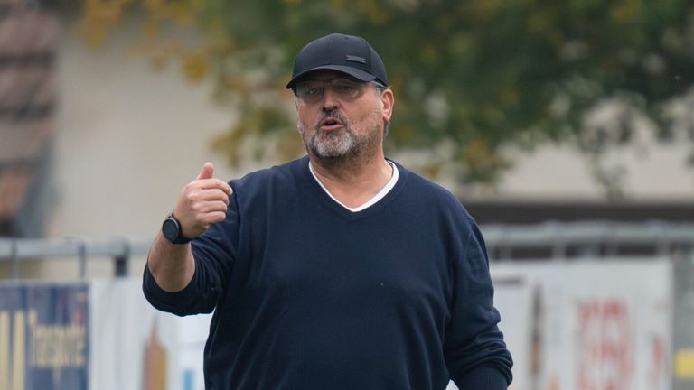 Ein Mann mit schwarzer Kappe und Brille zeigt den Daumen nach oben. Er trägt ein marineblaues V-Neck-Shirt und eine schwarze Armbanduhr.