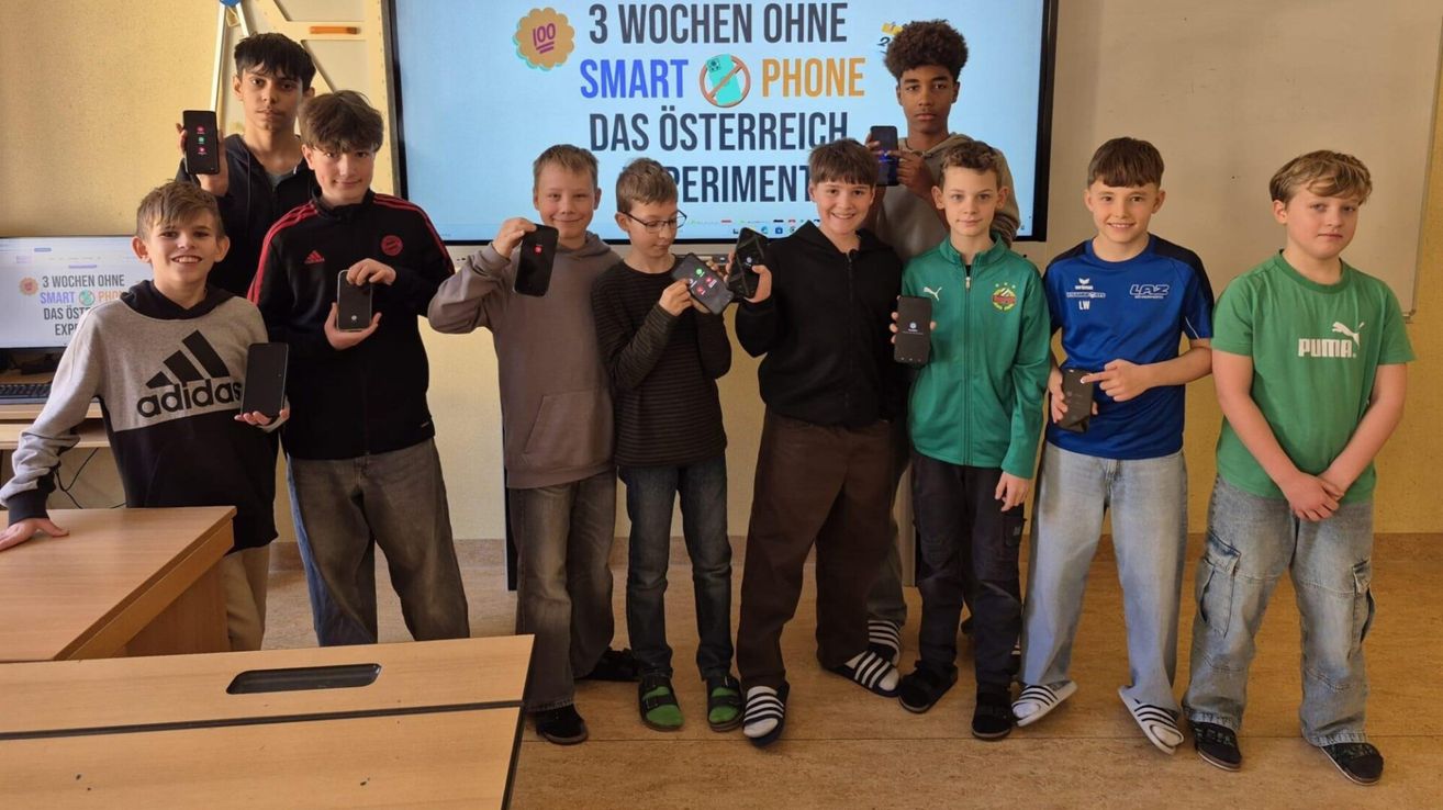 Eine Gruppe von Jungen steht in einem Klassenzimmer. Sie halten schwarze Geräte in den Händen. Ein Beamer mit Text steht dahinter.