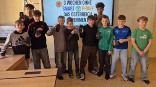 Eine Gruppe von Jungen steht in einem Klassenzimmer. Sie halten schwarze Geräte in den Händen. Ein Beamer mit Text steht dahinter.