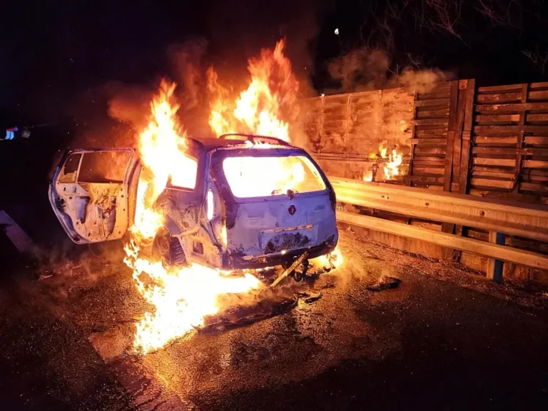 Ein blaues Auto steht mitten in der Nacht in Flammen am Straßenrand. Das Auto befindet sich in einem offenen Bereich, mit einer Holzbarriere dahinter. Das Feuer breitet sich aus, und das Auto ist fast vollständig verbrannt.