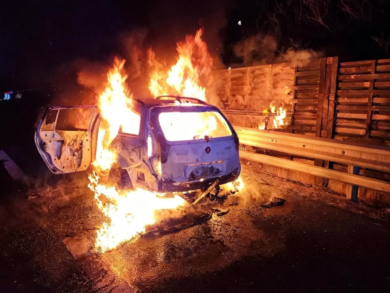 Ein blaues Auto steht mitten in der Nacht in Flammen am Straßenrand. Das Auto befindet sich in einem offenen Bereich, mit einer Holzbarriere dahinter. Das Feuer breitet sich aus, und das Auto ist fast vollständig verbrannt.
