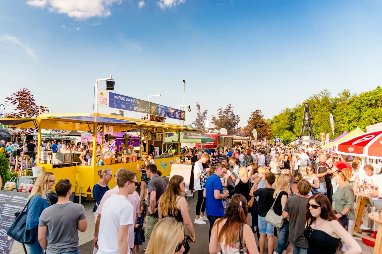 Eine große Menschenmenge versammelt sich bei einem Outdoor-Food-Festival unter einem klaren blauen Himmel. Verschiedene Essensstände sind aufgebaut, einer zeigt deutlich hausgemachtes Limoncello und Tee, mit Leuten, die sich anstellen, um zu kaufen.