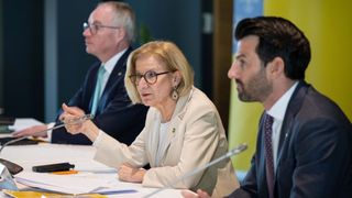 Drei Personen, zwei Männer und eine Frau, sitzen um einen Tisch herum bei einer Besprechung. Die Frau, die ein Mikrofon hält, scheint zu sprechen. Der Mann zu ihrer Rechten hört aufmerksam zu. Papiere und Stifte liegen auf dem Tisch.