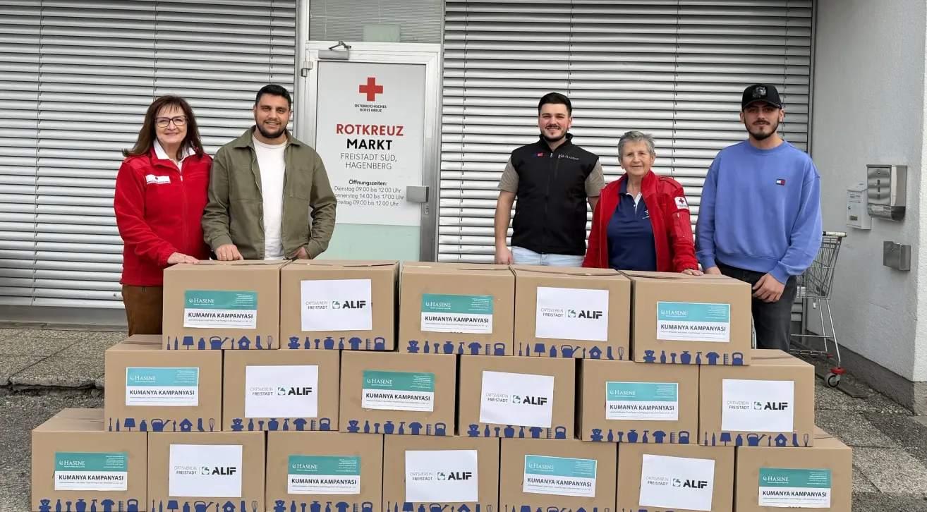Vier Personen stehen vor Kisten. Sie lächeln. Ein Schild hinter ihnen lautet 'Rotkreuz Markt Freistadt, Süd, Hagenberg'. Die Kisten tragen Aufkleber mit 'Kumanya Kampanyasi' und 'Hasene'.