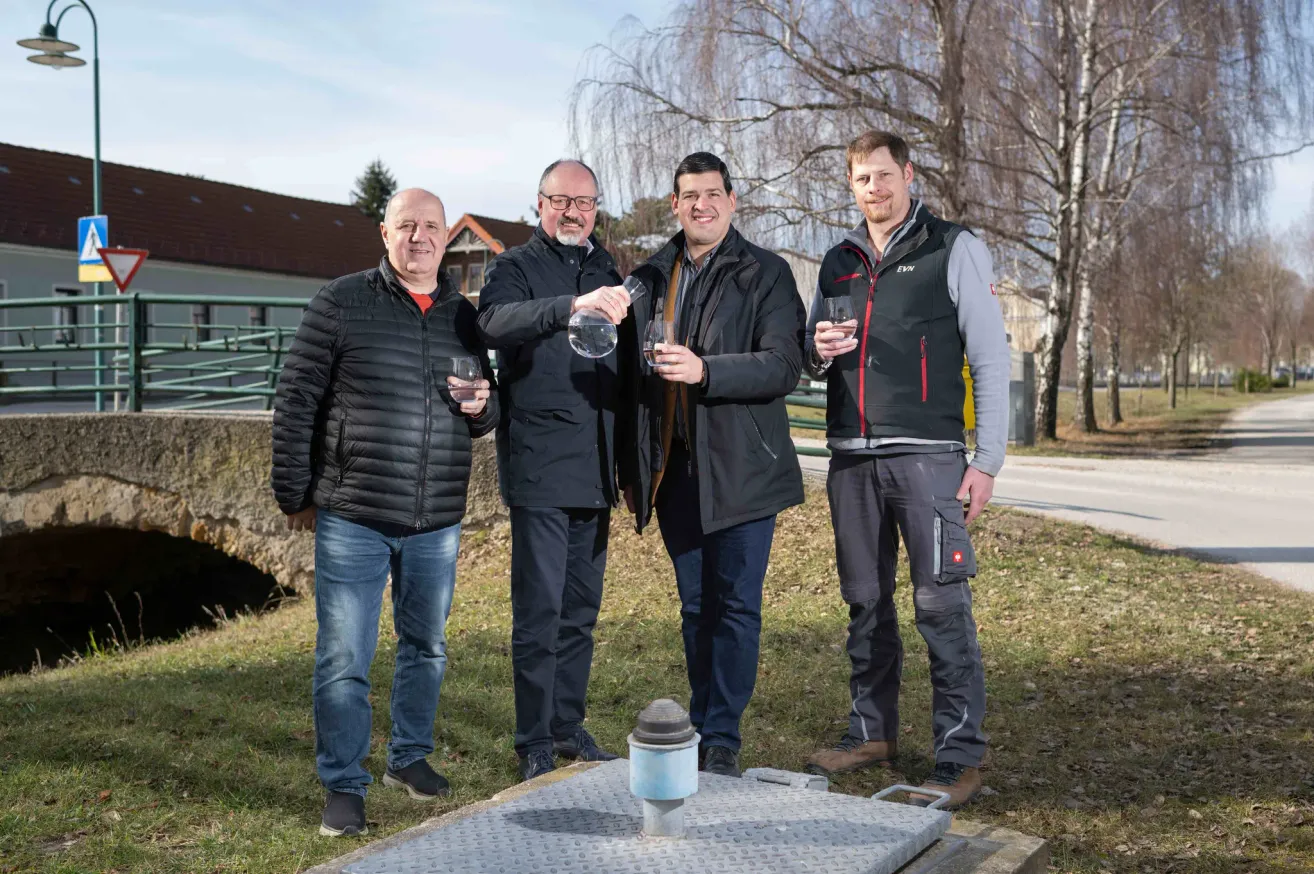 Vier Männer stehen auf einem Rasenfeld und halten Weingläser. Dahinter befindet sich eine Struktur mit einem Metalldeckel und einem zylindrischen Objekt. Bäume und ein Haus sind im Hintergrund zu sehen.