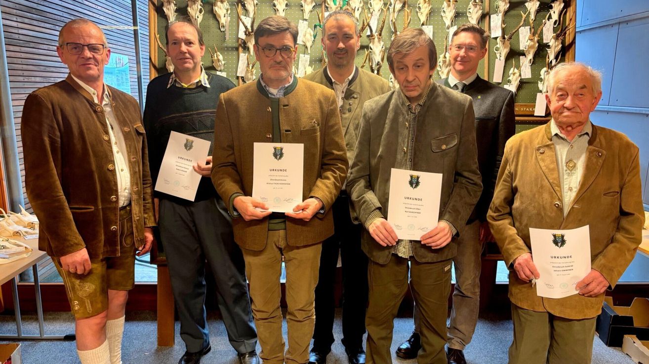 Eine Gruppe von Männern, gekleidet in Mänteln und Hosen, steht zusammen. Sie halten Urkunden und scheinen für ein Foto zu posieren. Die Männer stehen vor einer Wand mit verschiedenen Tierschädeln, die daran hängen.