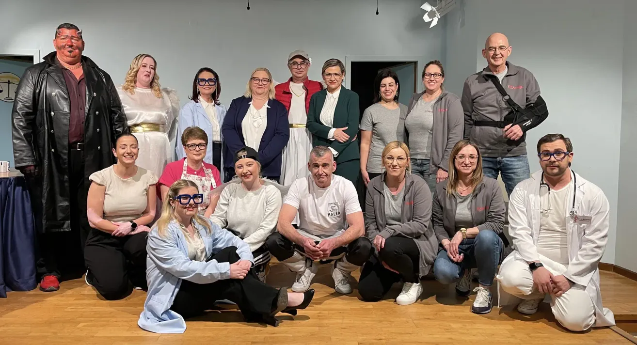 Eine Gruppe von Menschen posiert für ein Foto in einem Raum, einige stehend und andere sitzend. Sie tragen verschiedene Outfits und Accessoires.