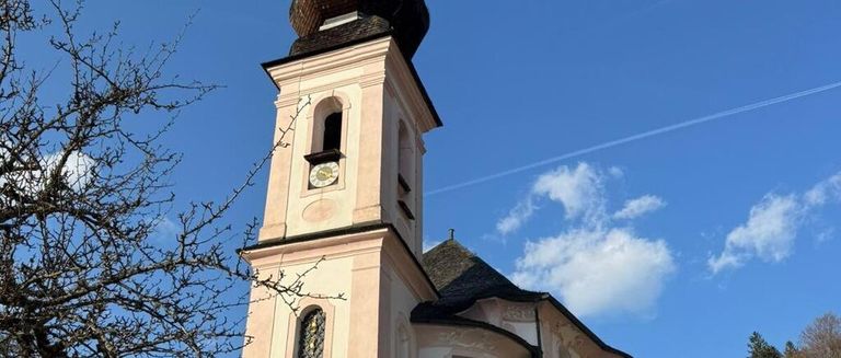 A church tower with a clock stands tall against a clear blue sky, featuring a steeple, stained glass windows, and a few bare branches.